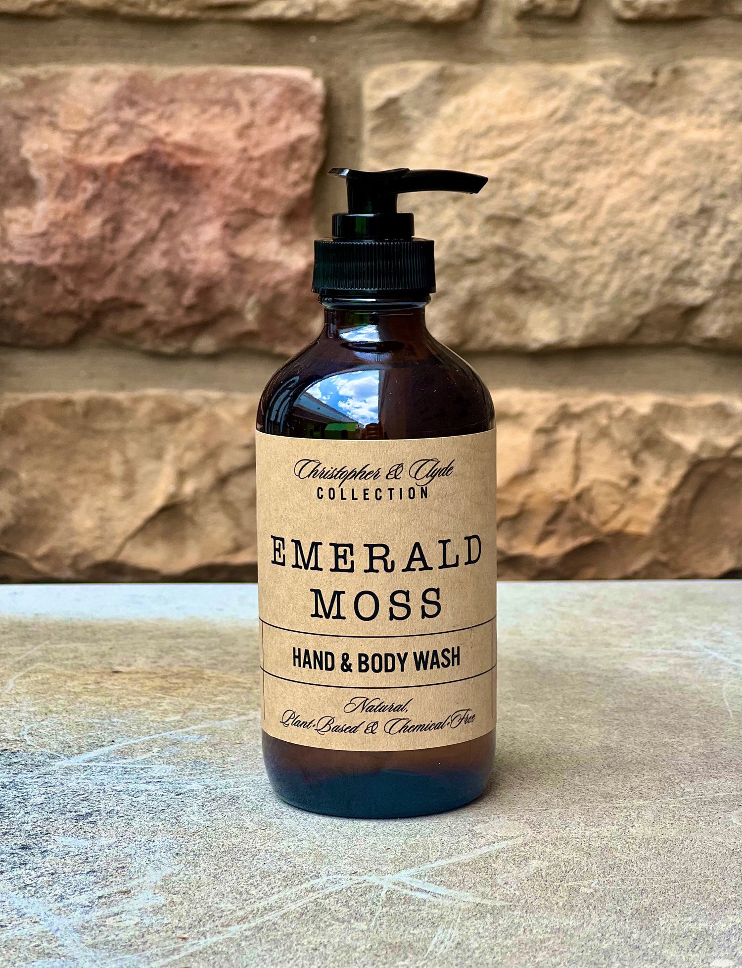 Hand and body wash bottle labeled 'Emerald Moss' with a stone wall background